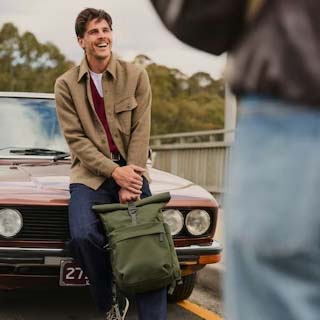 一名男子坐在一辆老爷车的棕色引擎盖上,手里拿着一个绿色天鹅绒老爷卷顶背包。另一个人站在前景中。 一名男子坐在一辆老爷车的棕色引擎盖上,手里拿着一个绿色天鹅绒老爷卷顶背包。另一个人站在前景中。